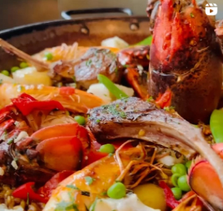 A close-up of a vibrant dish featuring lamb chops, large red crab claws, fresh vegetables, peas, and a mix of spices, served in a large pan.