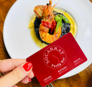 A red card for 'Socarrat Paella Bar' held by a person with red-painted nails, in front of a gourmet dish featuring black rice, shrimp, and garnish served on a white plate.